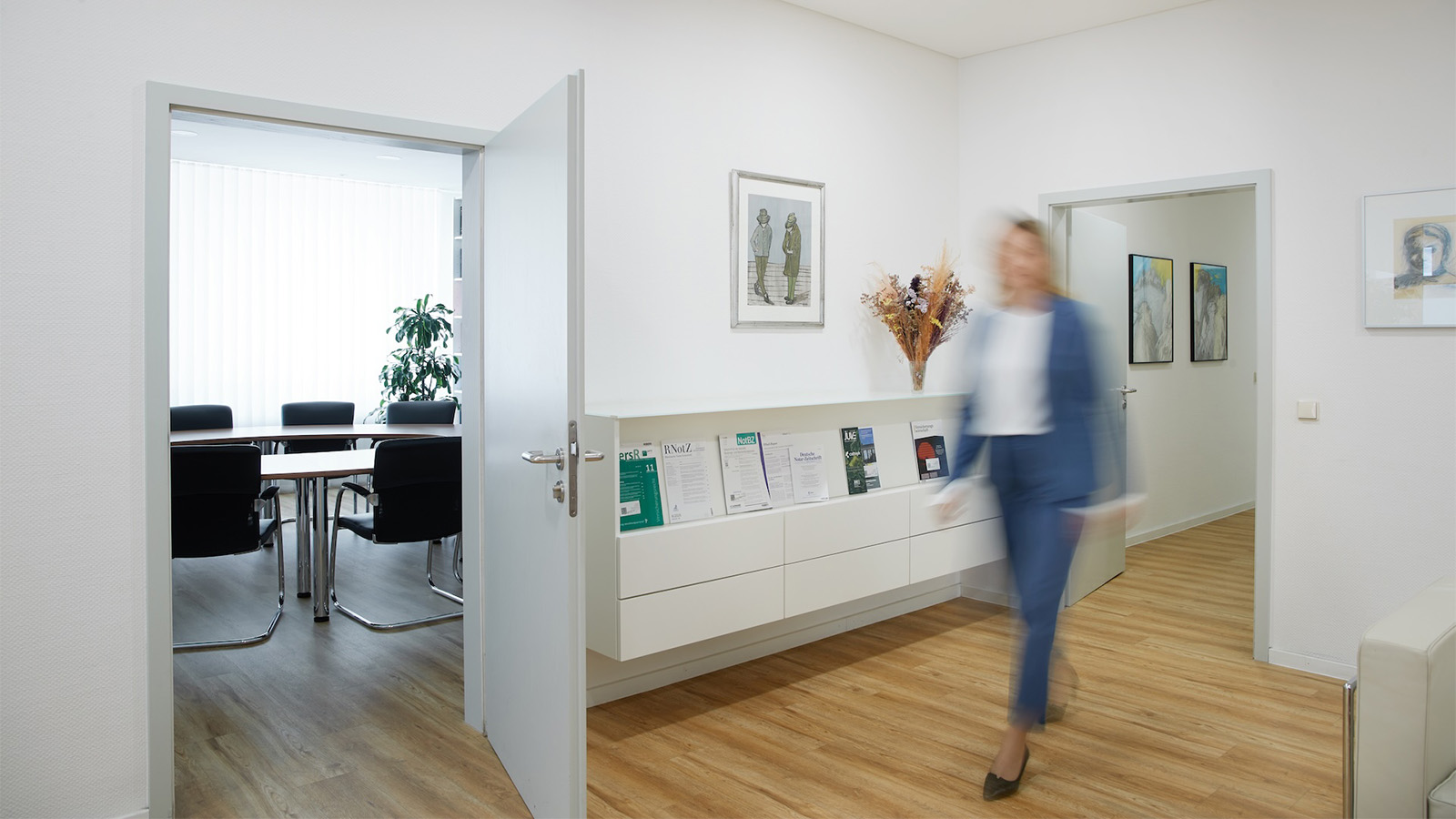 Eine Frau geht durch einen hellen Büroflur neben einem geöffneten Besprechungsraum.